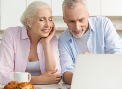 Image of happy mature loving couple family standing at the kitchen using laptop computer. Looking aside.