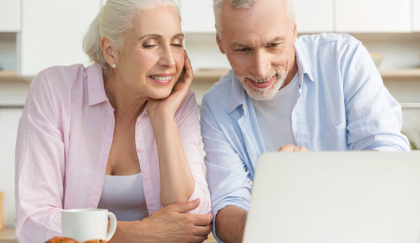 Image of happy mature loving couple family standing at the kitchen using laptop computer. Looking aside.